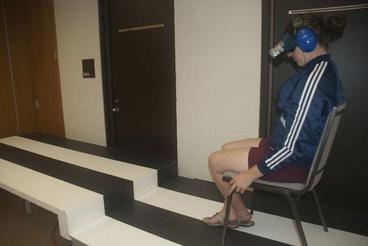 Photo of participant sitting on a platform wearing blur goggles and headphones, looking at a movable panel set to look like a step up. Both the floor and the panel have vertical black and white stripes.